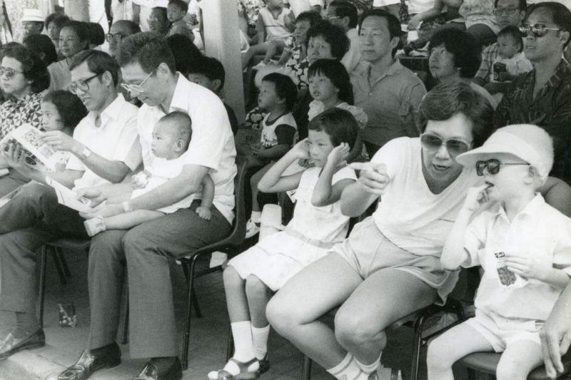 李显龙(抱着鸿毅)与何晶(右二)婚后,何晶为他再添两个儿子,图为夫妇带着原配所生的修齐和毅鹏到乌节路看妆艺大游行。坐在李显龙右手边的是前内阁部长李玉全。(档案照片)