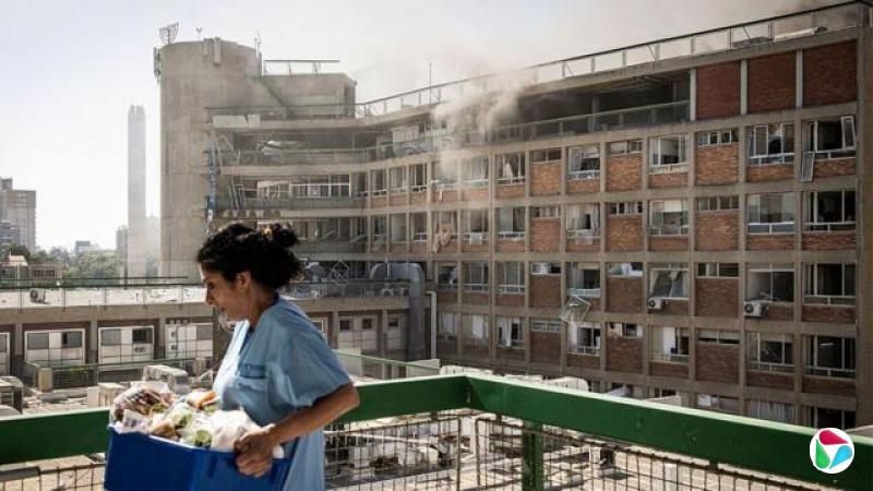 2025年6月19日,在以色列南部贝尔谢巴,伊朗导弹攻击后,索罗卡医院冒出浓烟,一名护士正提着医疗用品走过受损建筑物