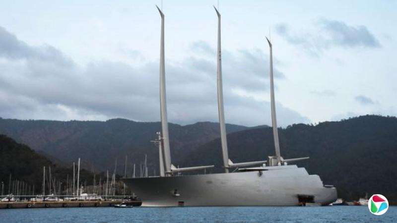 一艘外观引人注目的巨大银色超级游艇,带着高耸的桅杆,停泊在群山前方