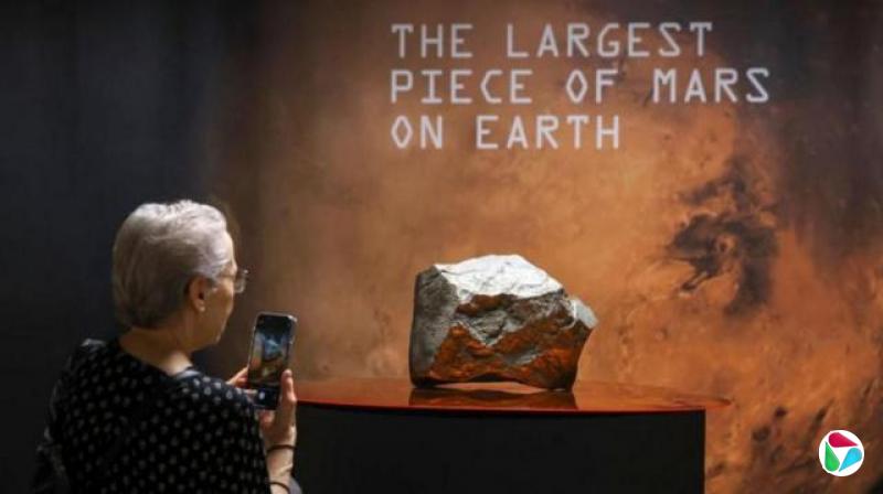 这颗在苏富比拍卖的最大火星陨石。旁边是拿着手机拍摄的一位白发、戴眼镜的女性。