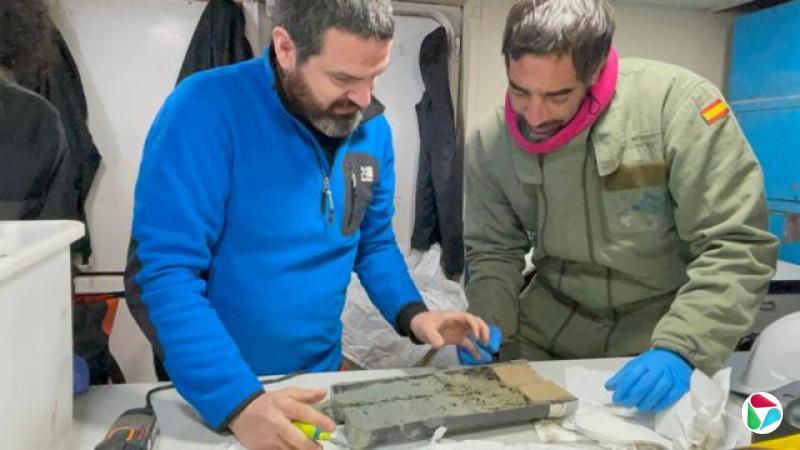 两位科学家站在桌旁,观察一根从南极海床收集到的绿色泥芯圆柱体。两人皆有深色头发和胡须;左侧研究人员穿着蓝色抓绒衣,右侧研究人员穿着绿色夹克和蓝色手套