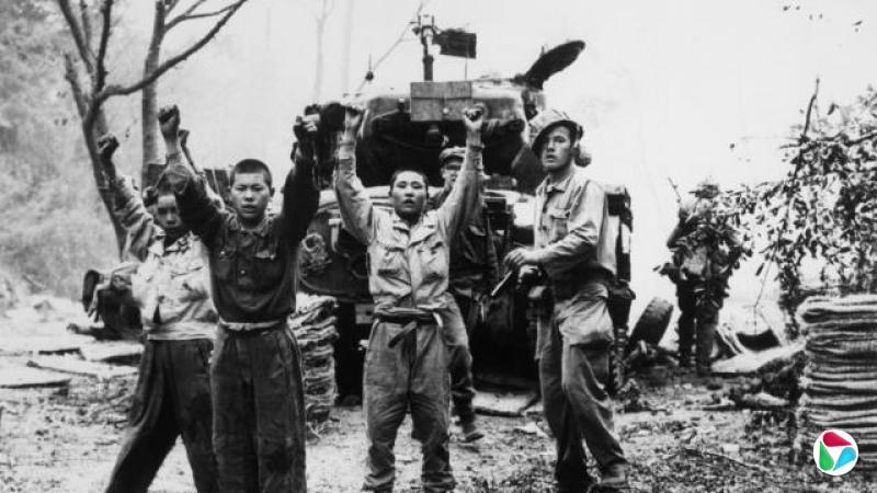 三名穿着军装的朝鲜男子举起双手,后方似乎是一名持枪的美国士兵,他的头盔歪斜地戴在头上。背景中有一辆坦克,还能隐约看到更多士兵的轮廓。这是一张黑白照片
