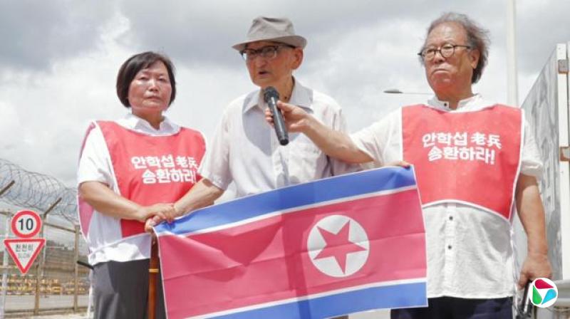 安学燮戴着眼镜、帽子和白衬衫,手持朝鲜国旗,身旁站着两位穿红色背心的志工——一人为他拿着麦克风,另一人握着他的右手