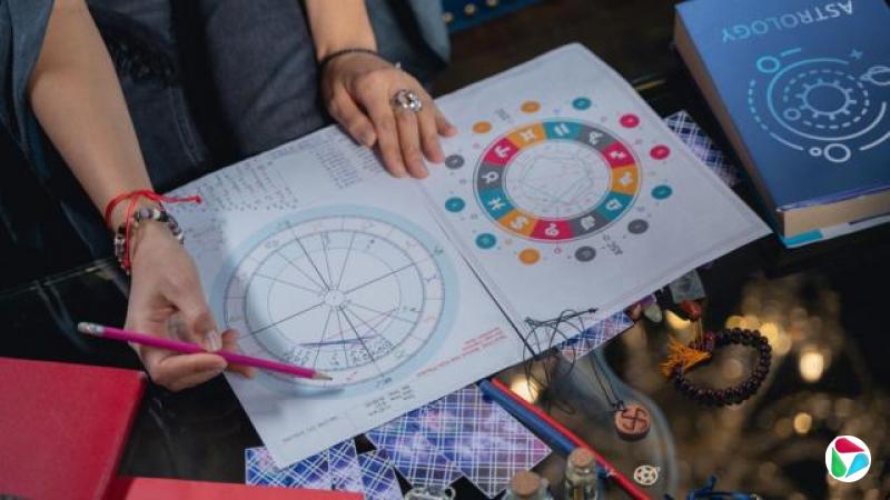 一张裁切照片显示一名女子的双手放在桌上,手持铅笔停在一张色彩缤纷的占星图上。桌面上散落着卡片、骰子,以及书本和铅笔。