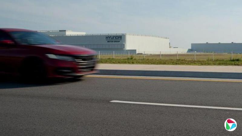 一辆红色轿车驶过位于乔治亚州的现代汽车工厂,该工厂是一座白色的建筑,周围有围墙及草地。