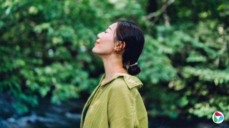 一位留着黑色长发并紮成低马尾的亚洲女性闭着眼睛仰望天空。她穿着一件绿色衬衫,背景是绿色的树木。
