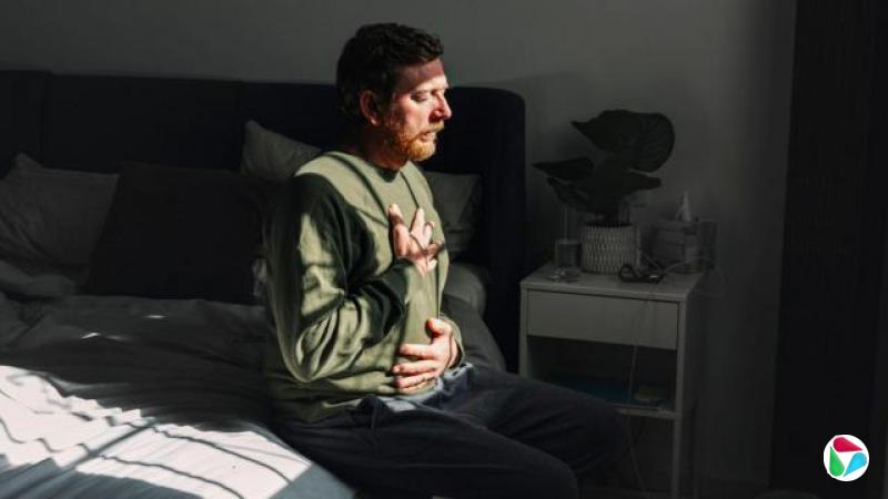 一个男人坐在床边,一只手放在胸口,另一只手放在腹部,窗外的阳光洒在他的脸上。