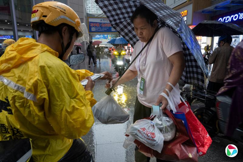 在赛格广场外,一名骑电动车的外卖骑手将餐食交给一位代送跑腿员,由他送到大楼里下单的顾客手中。