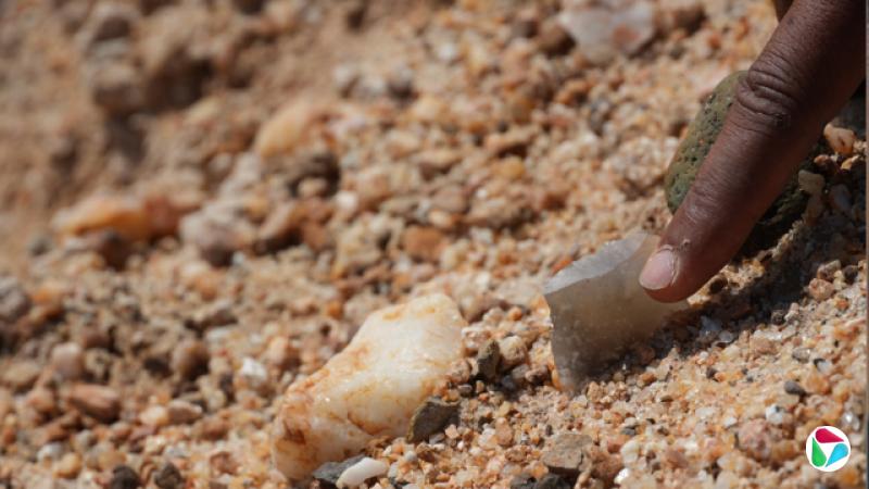 一只人的手正在指向或握住一件部分埋在沙质、碎石地面中的锐利石器。画面特写呈现出工具周围泥土与小石子的质地,焦点暗示这可能是在考古挖掘过程中发现或检查的一件潜在文物。