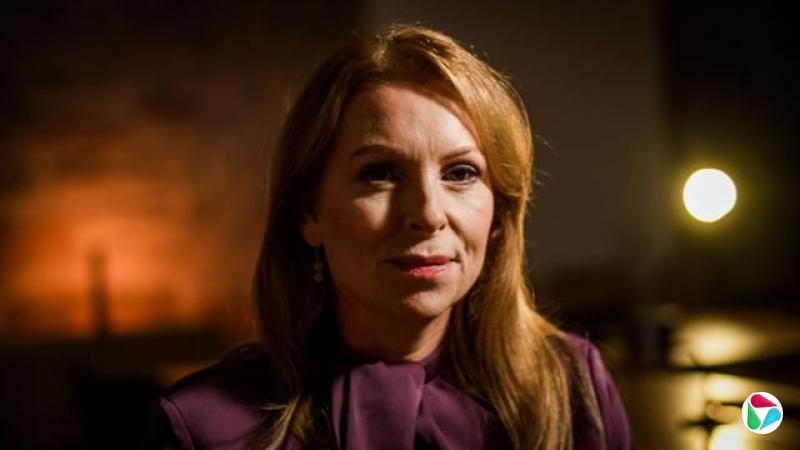 阿什·里根(Ash Regan)留着一头偏分的红色长发。她身穿一件深紫色的上衣,领口较高,胸前系着蝴蝶结。背景光线昏暗,色调温暖。一束光从她身后照过来,发出柔和的光芒,而背景的其余部分则模糊不清。