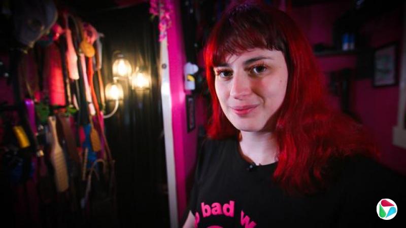 波塞琳·维多利亚正对着镜头微笑。她留着一头染成亮红色的长发,身穿一件印有亮粉色文字的黑色T恤。在左侧,可以看到挂钩上挂着一系列皮带和拍打板。房间光线昏暗。